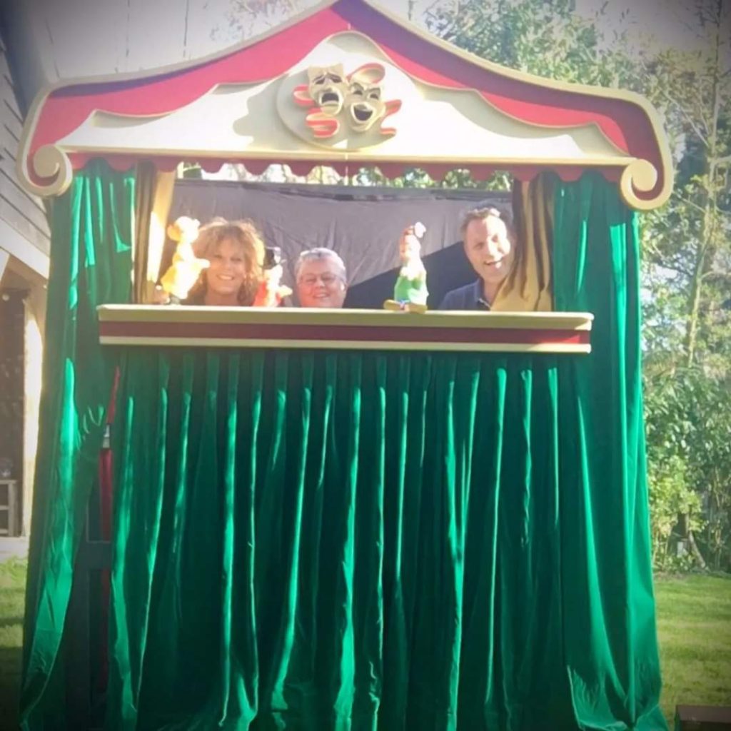 Drie spelers met handpoppen kijken lachend boven de rand van een poppenkast met groen fluwelen gordijn.