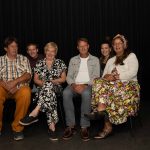 De zes spelers van Bedgeheimen naast elkaar op stoelen voor een zwart toneeldoek.
